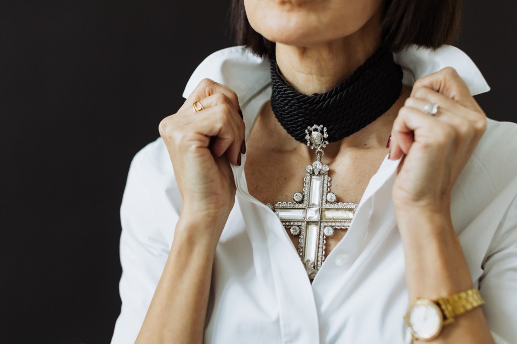 A close-up of a woman adjusting a Versace choker necklace made of silk rope and featuring a large glass cross, contrasted against her white collared shirt.